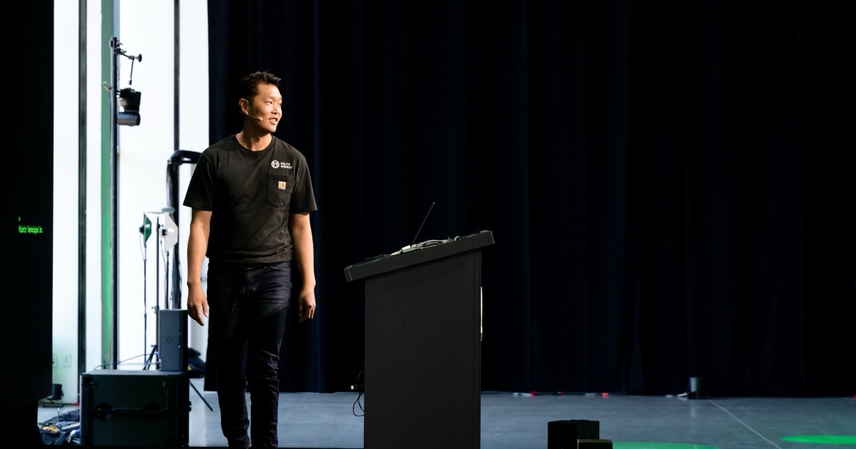 Frank Chen speaking beside a lectern on a black-curtain stage at Palantir DevCon, with green stage accents and no projected slide in frame.