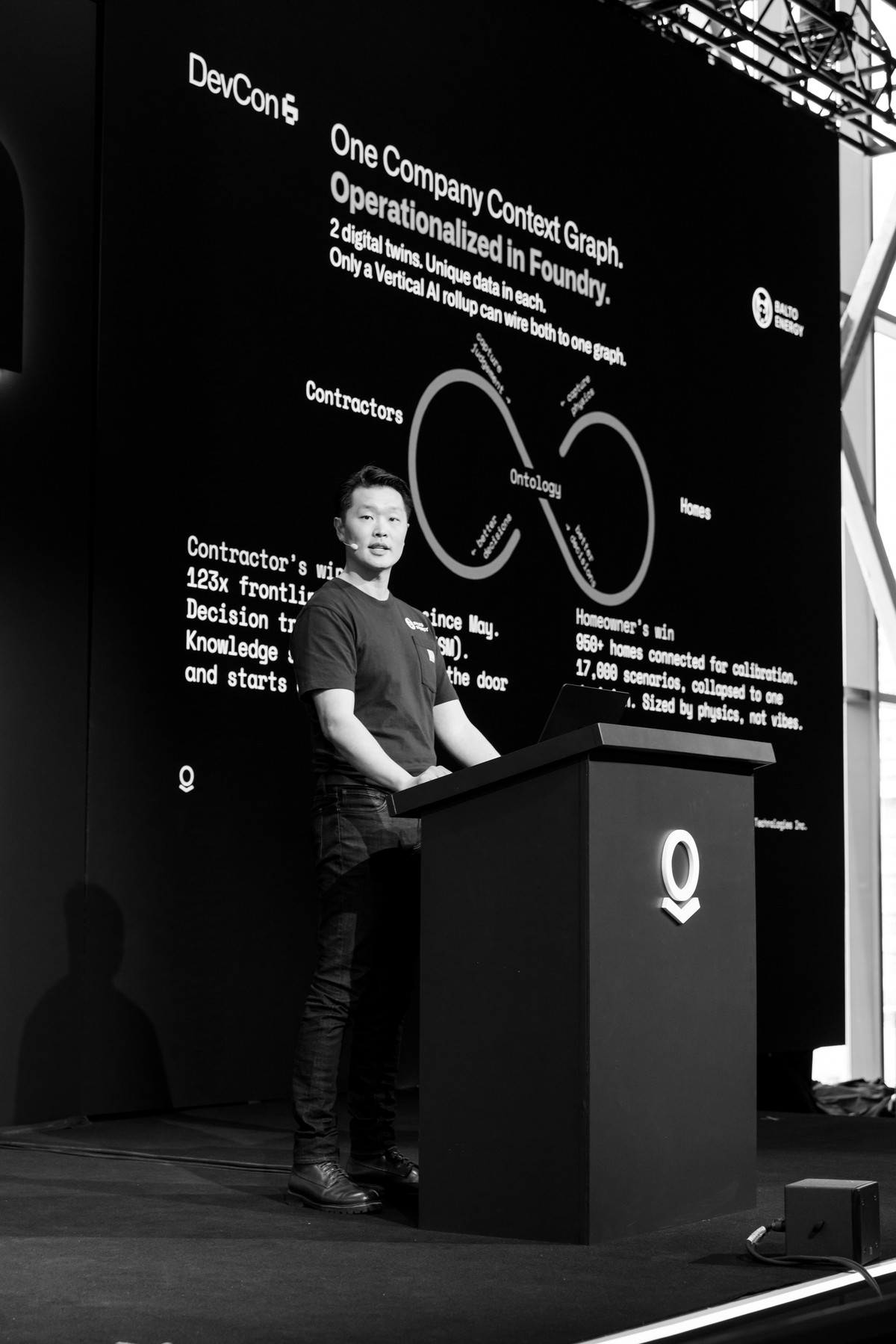 Frank Chen at a lectern on the Palantir DevCon stage, wearing a headset microphone, in front of a screen showing a company context graph linking contractors and homes through a shared ontology.