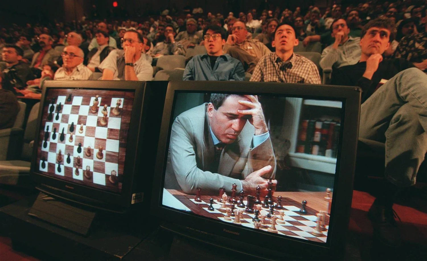 Garry Kasparov at the board during the 1997 Deep Blue match, head in hand, shown on television monitors in front of a packed auditorium.