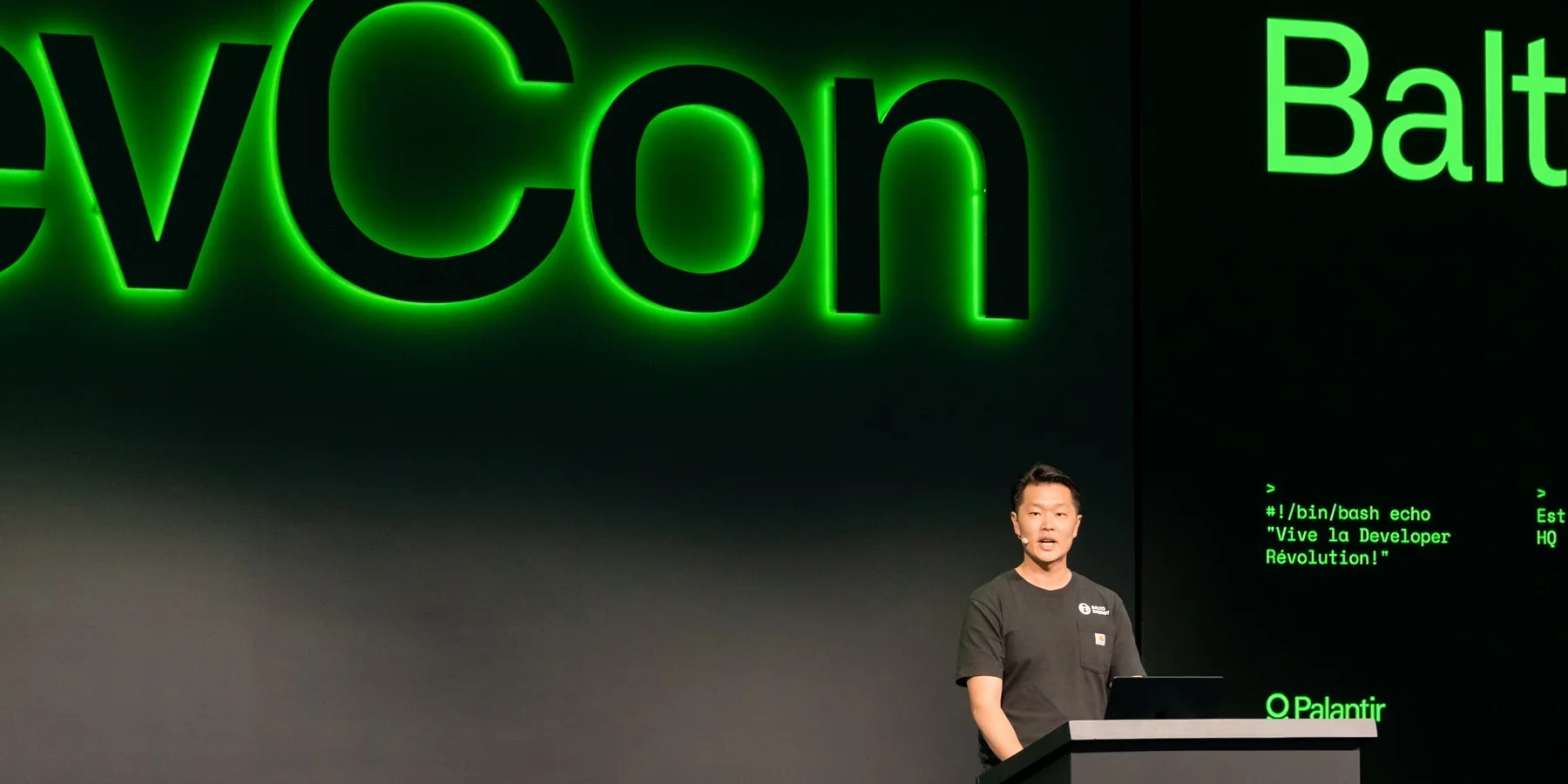 Frank Chen at a lectern on the DevCon stage, the illuminated DevCon wordmark filling the wall behind him and his title slide lit to the right.